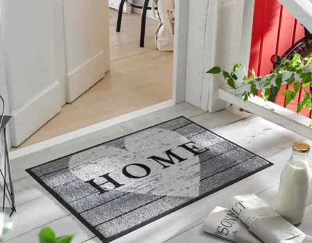 A stylish gray and black patterned home mat inside a large white heart placed on a light wooden entryway deck outside a door A stylish gray and black patterned home mat inside a large white heart placed on a light wooden entryway deck outside a door