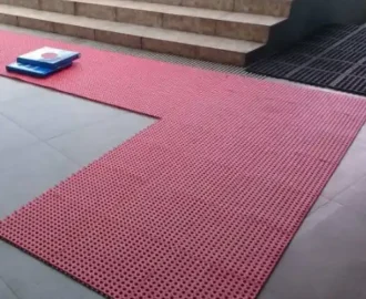 L-shaped section of red rubber ablution mats laid out on a gray tiled floor, extending from the bottom of a light brown or beige set of exterior steps. L-shaped section of red rubber ablution mats laid out on a gray tiled floor, extending from the bottom of a light brown or beige set of exterior steps.