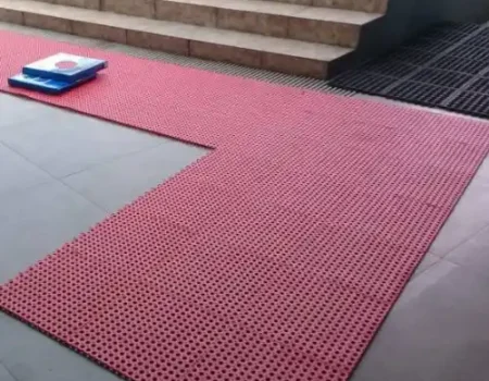L-shaped section of red rubber ablution mats laid out on a gray tiled floor, extending from the bottom of a light brown or beige set of exterior steps. L-shaped section of red rubber ablution mats laid out on a gray tiled floor, extending from the bottom of a light brown or beige set of exterior steps.